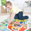0000772504164-1 A young boy in a yellow shirt and jeans assembles the Melissa & Doug Ms. Rachel Floor Puzzle Farm Ms Rachel while kneeling on a round rug in a bright, tidy room.