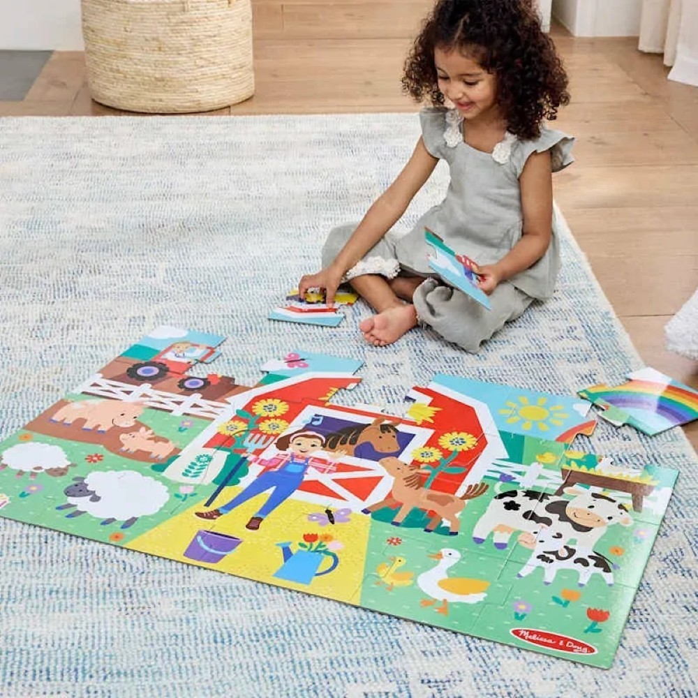 0000772504164-3 A child in a gray dress sits on a rug, assembling the Melissa & Doug Ms. Rachel Floor Puzzle Farm, which features colorful images of animals, a barn, and a farmer.