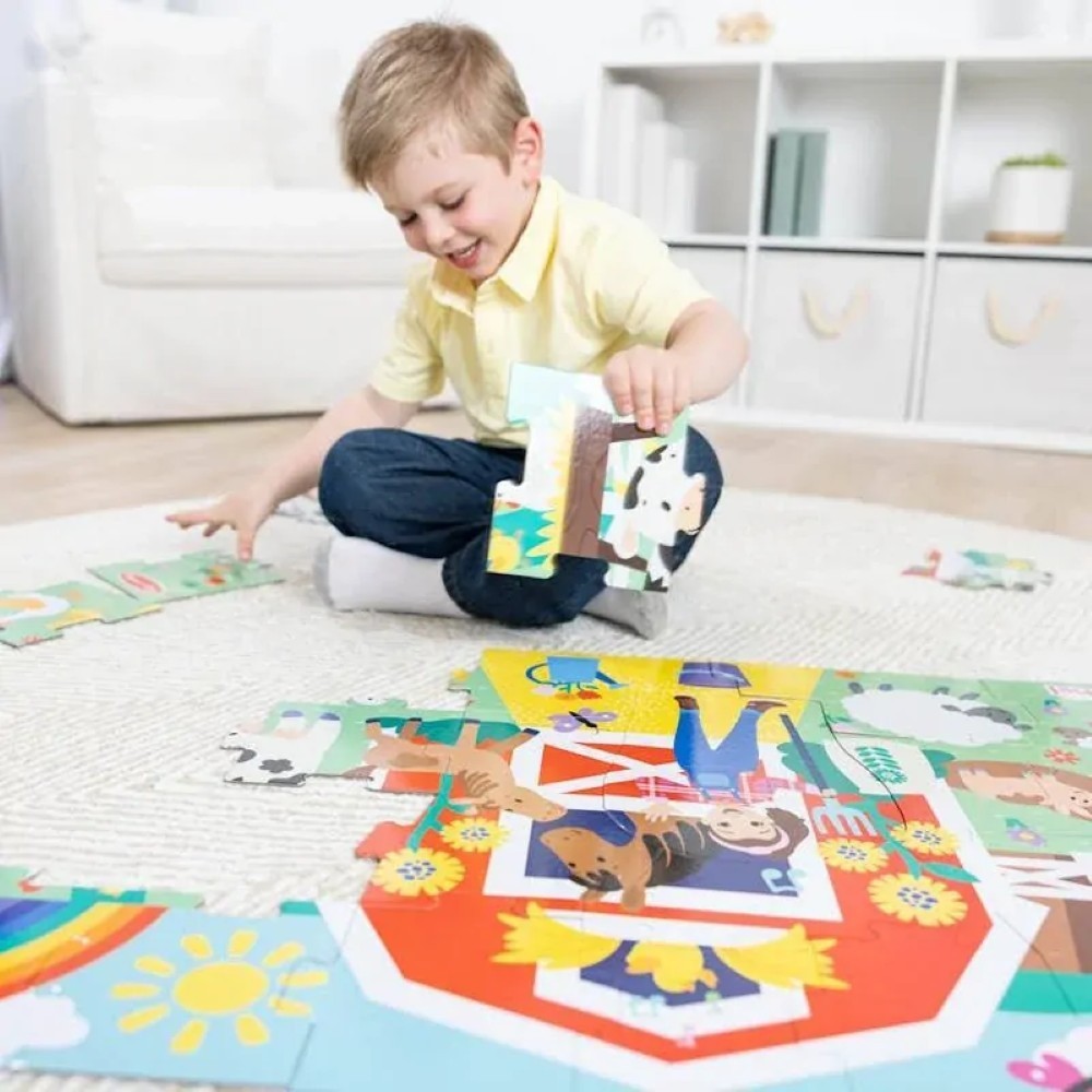 0000772504164-7 A young child sits on a rug in a bright, tidy room, assembling the Melissa & Doug Ms. Rachel Floor Puzzle Farm, featuring large, colorful pieces with various animals and a barn.