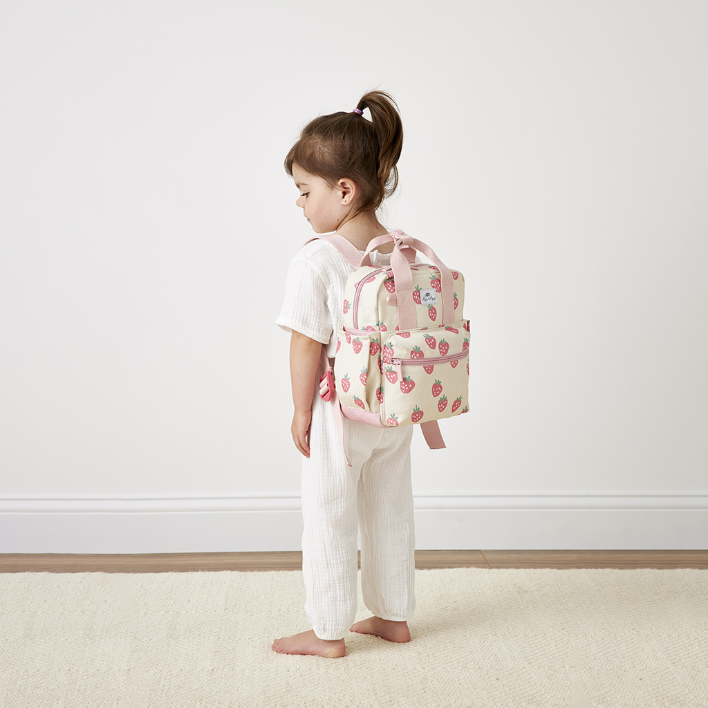 A young child with light brown hair stands in profile, clad in an all-white outfit and carrying a pink strawberry-patterned backpack. She is barefoot on a light-colored rug against a white wall, with the Itzy Ritzy Sweetie Strap Plus Bear Silicone Pacifier Clip clipped to her attire.