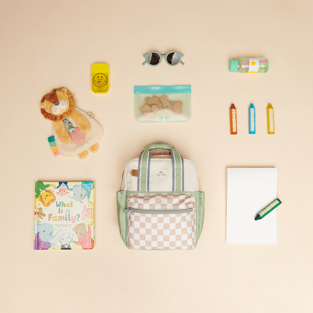 Top view of a neatly arranged set of items including a checkered backpack, stuffed lion toy, children's book, juice bottle, toy blocks, ziploc bag with cookies, sunglasses, notebook, green marker, and an Itzy Ritzy Sweetie Strap Plus Bear Silicone Pacifier Clip.