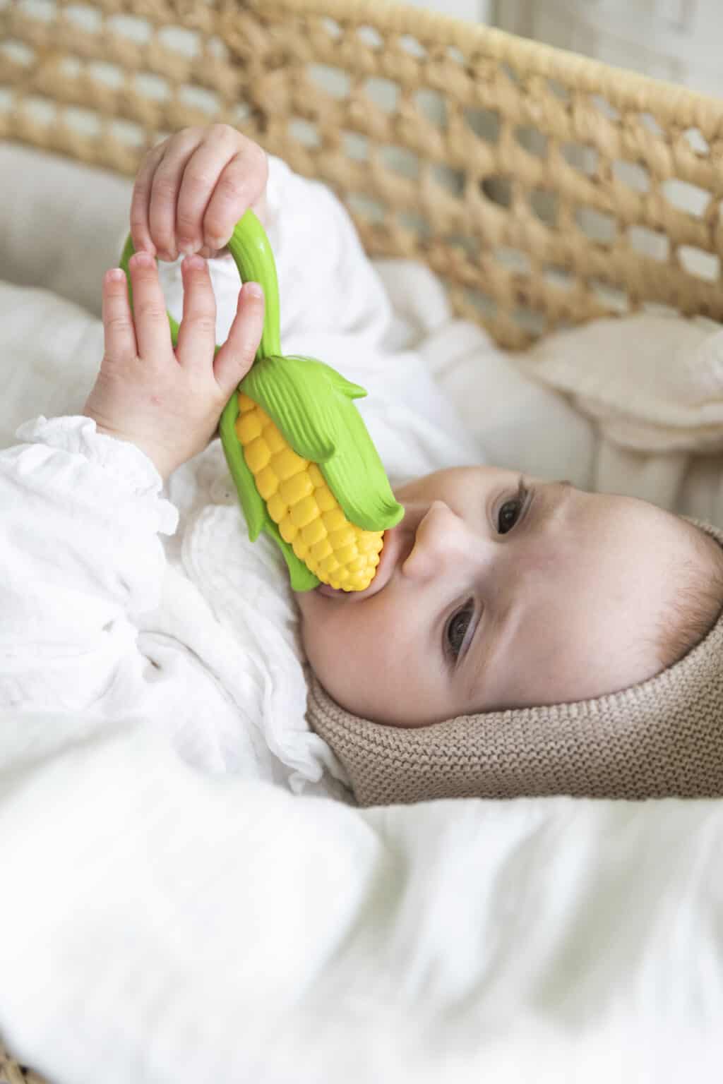 A baby is chewing on an Oli & Carol Corn Rattle Toy Teether Baby Natural Rubber in a basket.