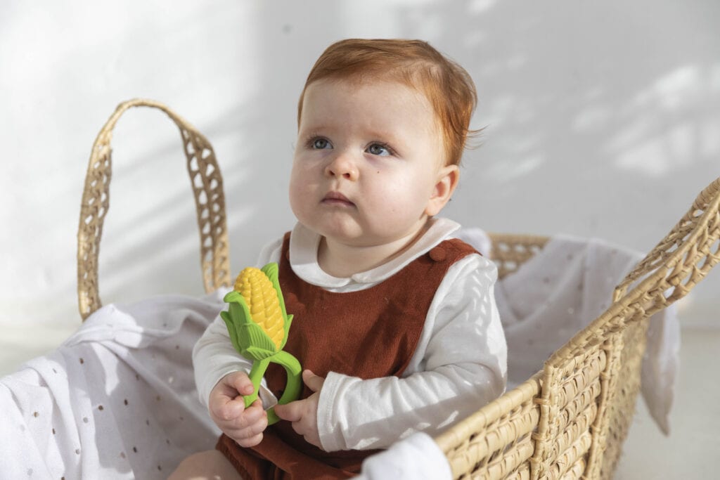 A baby is sitting in a basket holding an Oli & Carol Corn Rattle Toy Teether Baby Natural Rubber.