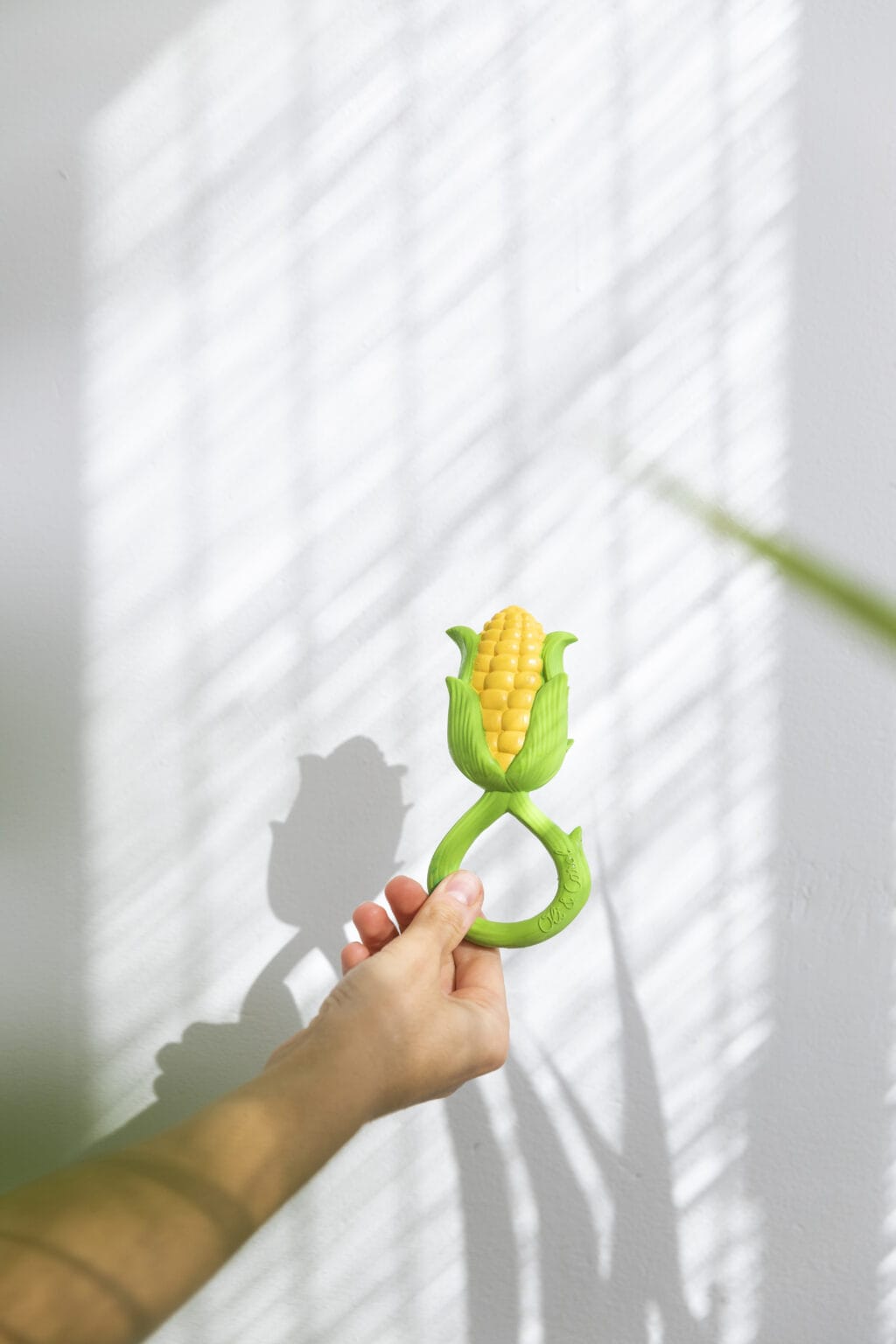 A person holding an Oli & Carol Corn Rattle Toy Teether Baby Natural Rubber in front of a window.