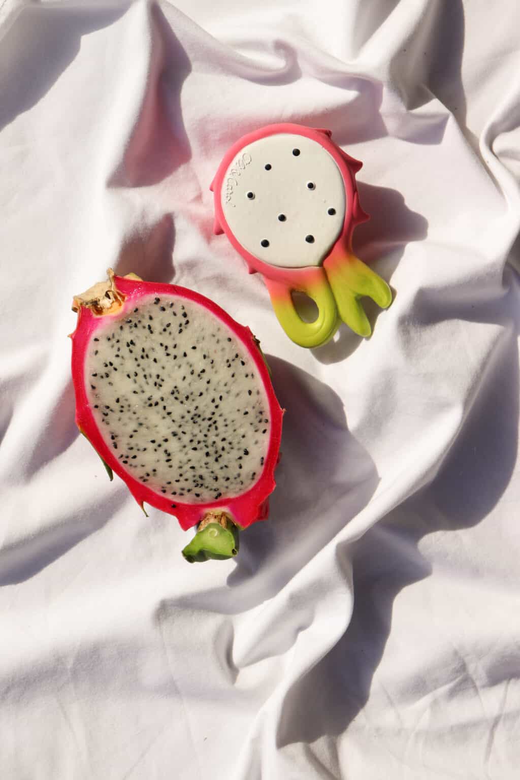 Two Oli & Carol Fucsia de Dragonfruit Baby Teether Natural Rubber shaped plates on a white cloth.