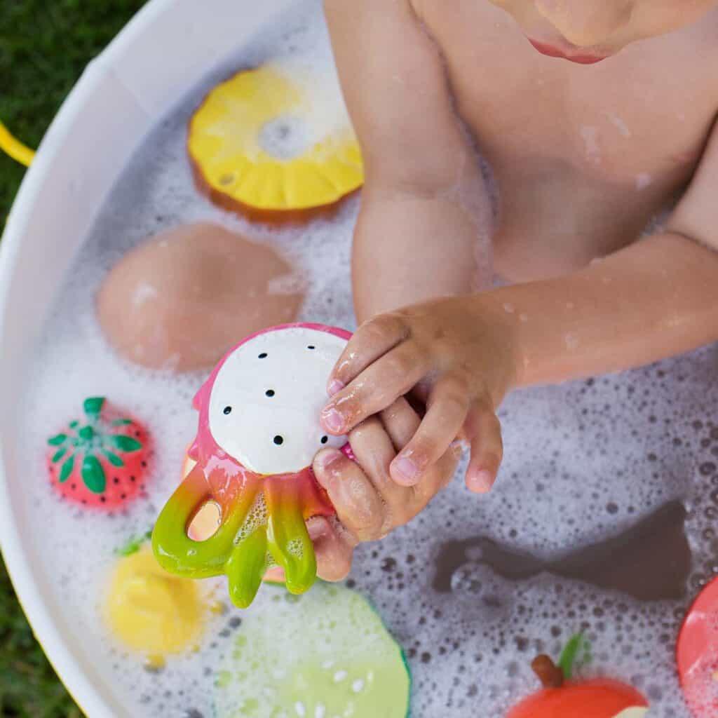 A child playing with the Oli & Carol Fucsia de Dragonfruit Baby Teether Natural Rubber in a tub.