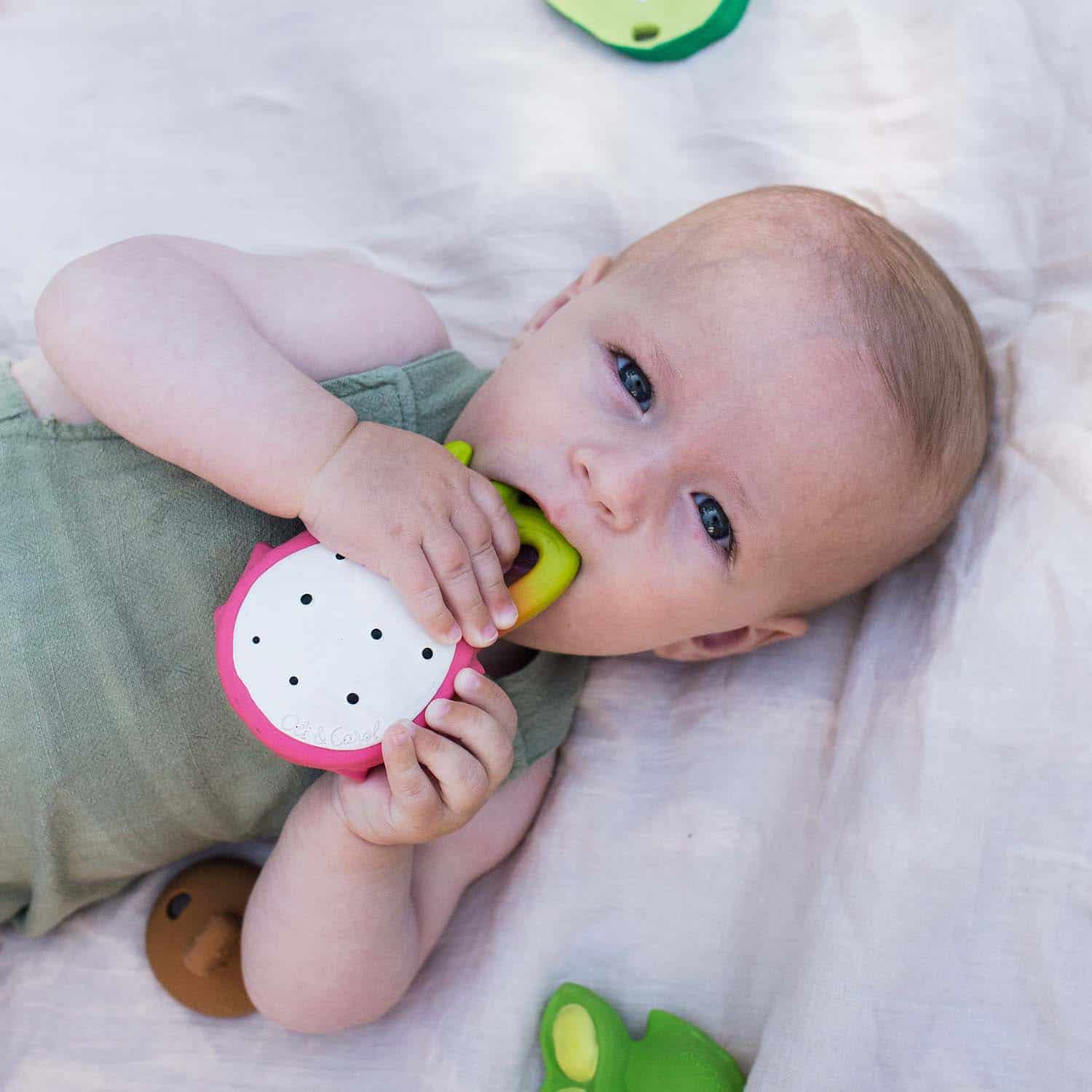 Oli & Carol Fucsia de Dragonfruit Baby Teether Natural Rubber (3) A baby is laying on a blanket with the Oli & Carol Fucsia de Dragonfruit Baby Teether Natural Rubber.
