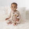 A baby is sitting on a white blanket with an Itzy Ritzy Soothe & Store Pacifier Clip & Storage Pouch.
