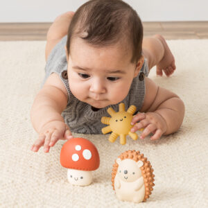 Baby lying on stomach reaching for the Itzy Ritzy Sensory Blocks 10-Piece Set with a plush sun and hedgehog nearby on a carpeted floor.