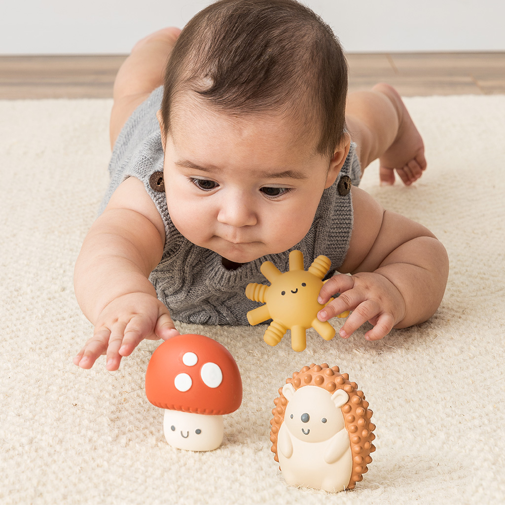 BL853201_Itzy_Ritzy_Toy_Itzy_Blocks_DigitalShelf Baby lying on stomach reaching for the Itzy Ritzy Sensory Blocks 10-Piece Set with a plush sun and hedgehog nearby on a carpeted floor.