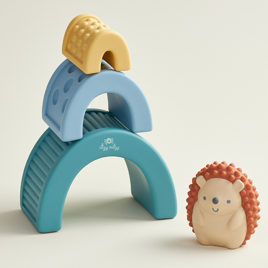 BL853201_Itzy_Ritzy_Toy_Itzy_Blocks_DigitalShelf A colorful stack of Itzy Ritzy Sensory Blocks 10-Piece Set featuring a blue arch and a yellow piece on top, accompanied by a smiling hedgehog-shaped toy, on a neutral background.