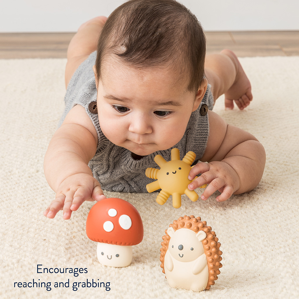 BL853201_Itzy_Ritzy_Toy_Itzy_Blocks_DigitalShelf Infant lying on stomach on a carpet, reaching towards an Itzy Ritzy Sensory Blocks 10-Piece Set and a toy hedgehog.