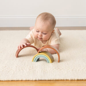 Baby lying on a rug playing with a wooden rainbow toy.