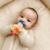 An infant in a beige outfit lies on a cream-colored surface, holding and chewing on a colorful teething toy.