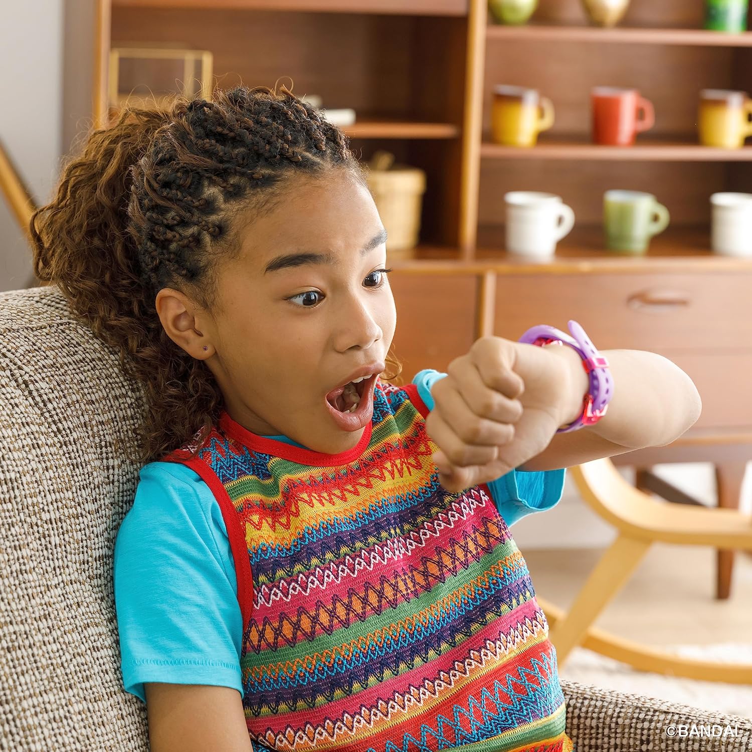 A young child with braided hair looks surprised while glancing at their wrist, which has a colorful watch. The child is indoors, sitting in a living room with shelves and colorful mugs in the background.