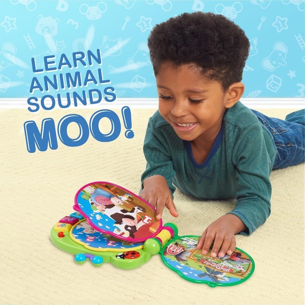 cocomelon-learningbook-103 A young boy lies on a carpet, smiling as he plays with the CoComelon Learning Book Interactive Toy. The text on the wall reads “LEARN ANIMAL SOUNDS MOO!” against a blue patterned background with playful icons.