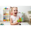 cocomelon-singingremote-71 A young child with pigtails smiles and holds the CoComelon Learn and Play Singing Remote Toddler Musical Toy Microphone, surrounded by shelves and toys in a cheerful playroom.