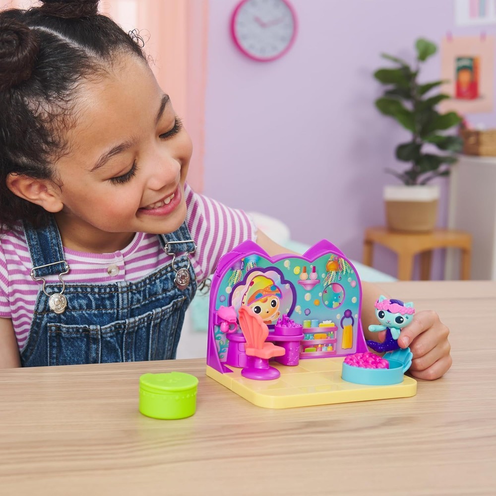 gabbydollhouse-mercatsparoom-82 A young girl in overalls plays with the Gabby’s Dollhouse MerCat’s Spa Room Playset at a table in a brightly decorated room with pastel walls and plants.