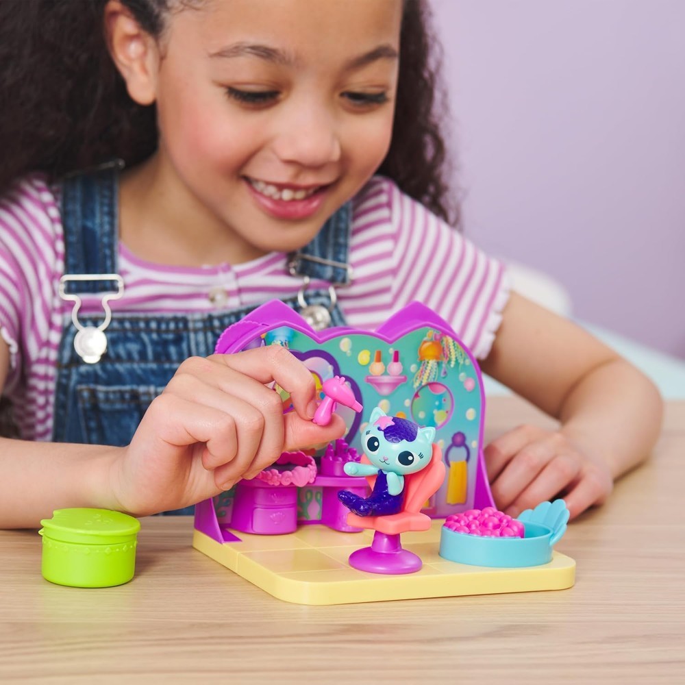 gabbydollhouse-mercatsparoom-83 A child in overalls plays with the Gabby’s Dollhouse MerCat’s Spa Room Playset on a wooden table, enjoying the small blue MerCat figure and colorful accessories nearby.