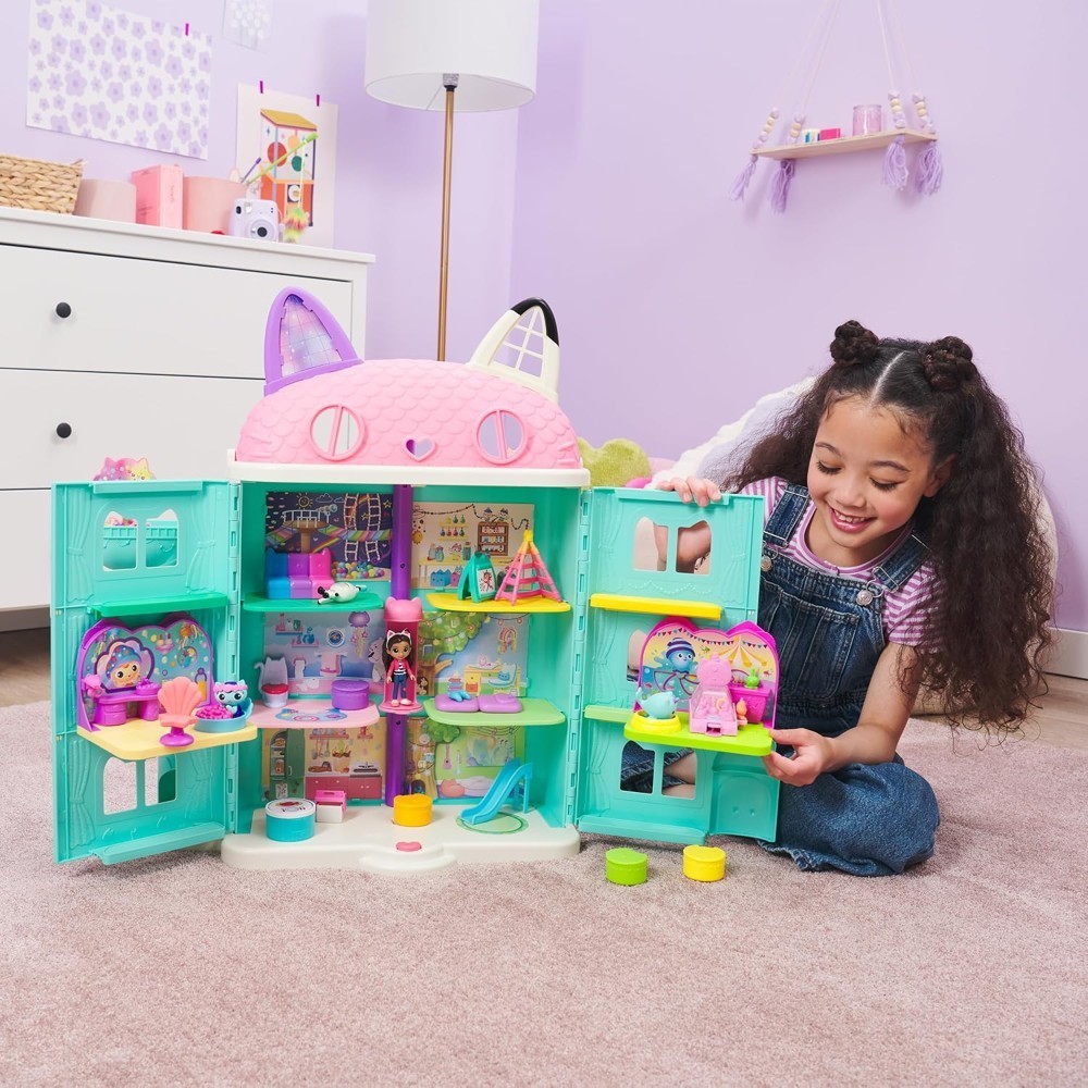 gabbydollhouse-mercatsparoom-86 A young girl smiles as she plays with the Gabby’s Dollhouse MerCat’s Spa Room Playset, which features a colorful, multi-story dollhouse with cat ears and includes several small dolls like the MerCat figure along with surprise toy furniture inspired by the Netflix series. She holds a doll in her right hand while enjoying the imaginative play set.