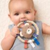 itzy-ring-pal-bear-02 Baby lying on a soft surface, holding and chewing on a circular teething toy with a plush bear and colorful ribbons.