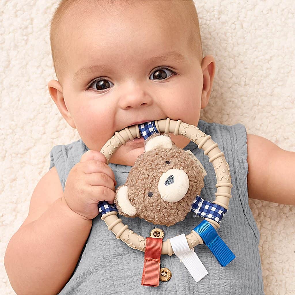 itzy-ring-pal-bear-02 Baby lying on a soft surface, holding and chewing on a circular teething toy with a plush bear and colorful ribbons.