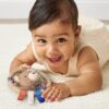 itzy-ring-pal-bear-03 A baby wearing a cream outfit lies on a light rug, smiling while holding a plush bear toy with ribbon tags.