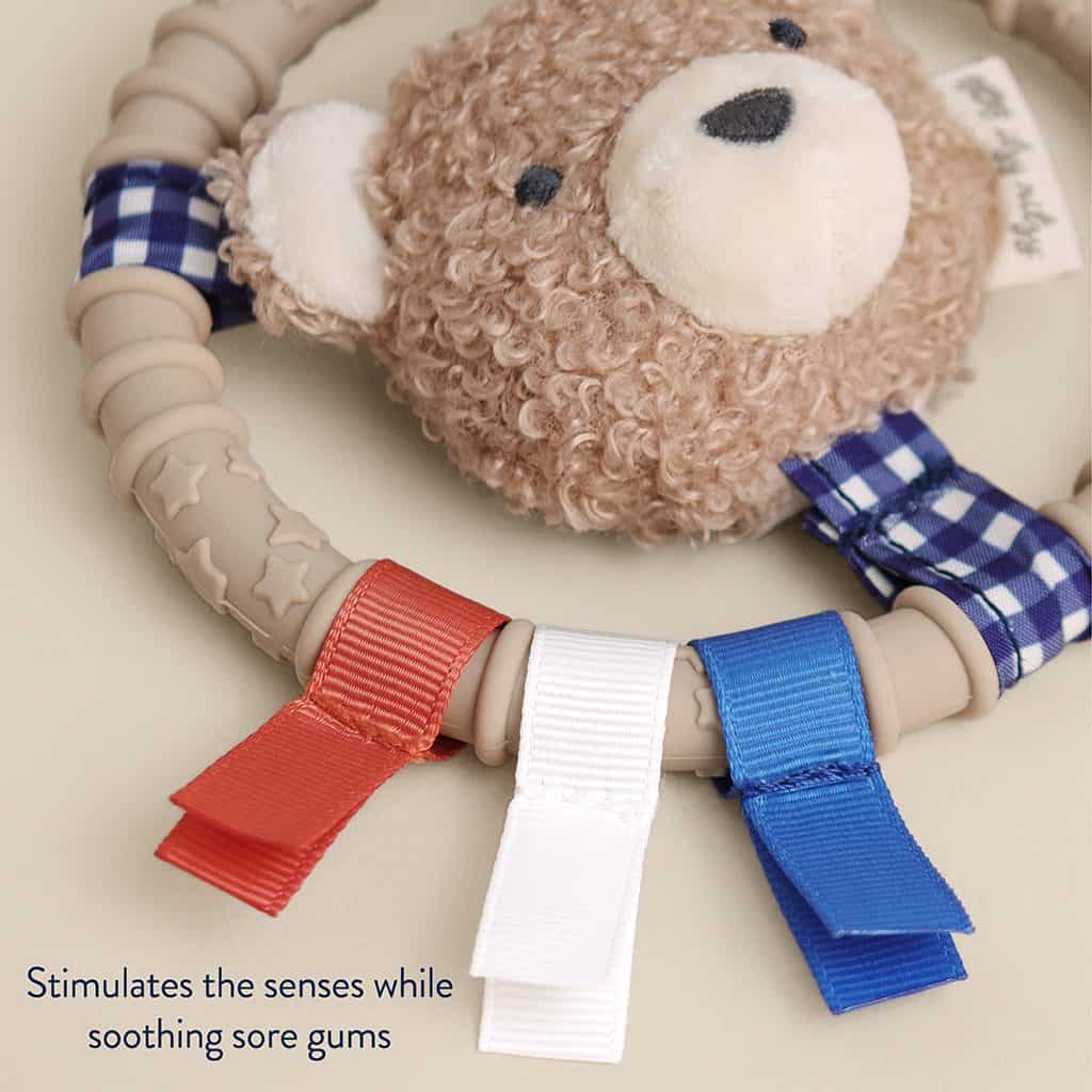 itzy-ring-pal-bear-04 Plush teddy bear teething ring with textured beige ring and red, white, and blue ribbon tags. Text reads: “Stimulates the senses while soothing sore gums.”.