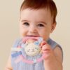 itzy-ring-pal-kitty-02 Baby in a light purple outfit holds a pink ring toy with a cat face, fabric tags, and blue accents, against a neutral background.