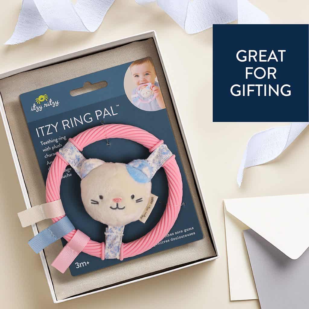 itzy-ring-pal-kitty-04 Gift box with a pink Itzy Ritzy “Ring Pal” teething ring featuring a plush cat face, displayed next to gift wrapping materials and a card that says “Great for Gifting.”.