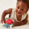 itzy-ring-pal-tomato-02 A baby in a sleeveless cream outfit lies on a soft surface, smiling and playing with a plush red toy that has colorful fabric tags.
