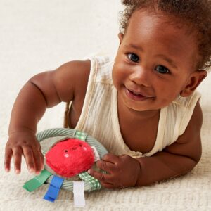 A baby in a sleeveless cream outfit lies on a soft surface, smiling and playing with a plush red toy that has colorful fabric tags.