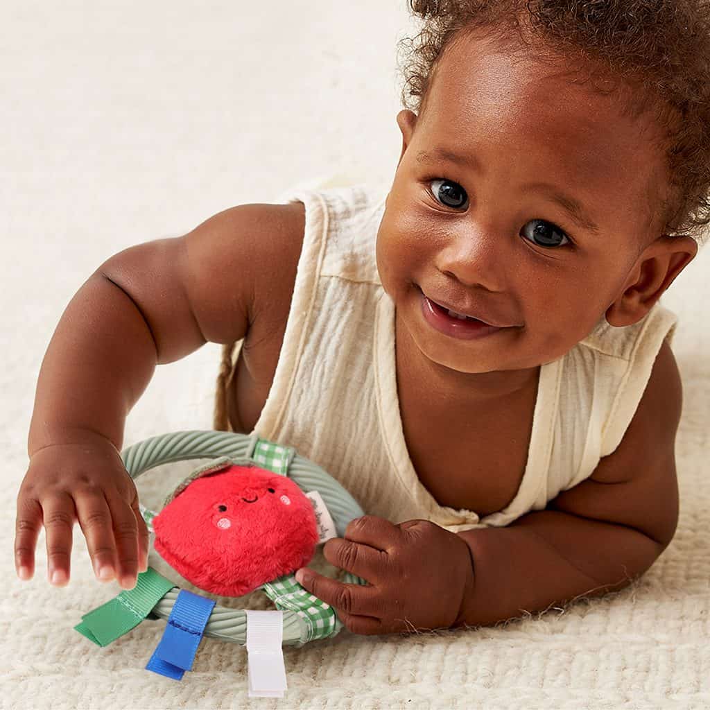 itzy-ring-pal-tomato-02 A baby in a sleeveless cream outfit lies on a soft surface, smiling and playing with a plush red toy that has colorful fabric tags.