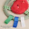 itzy-ring-pal-tomato-03 Gray teething ring with green, blue, and white ribbon tags and a plush red smiling face attached. Text reads “Stimulates the senses while soothing sore gums.”.