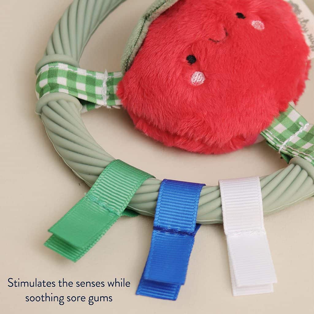itzy-ring-pal-tomato-03 Gray teething ring with green, blue, and white ribbon tags and a plush red smiling face attached. Text reads “Stimulates the senses while soothing sore gums.”.