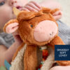 A baby holding a brown and orange cow-themed stuffed toy with a label reading "Snuggly Soft Lovey.