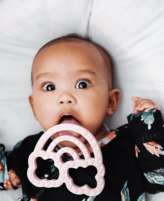 A baby is laying on a bed with a toy in her mouth.