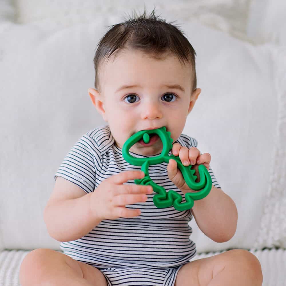 A baby is sitting on a bed with a green toy in his mouth.
