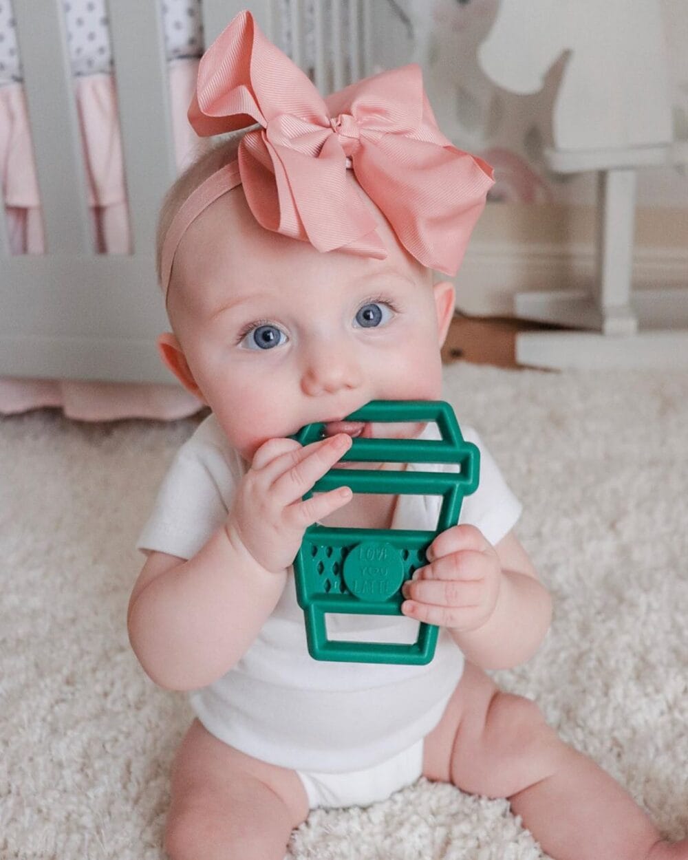 A baby is sitting on the floor with a Chew Crew Silicone Baby Teether in her mouth.