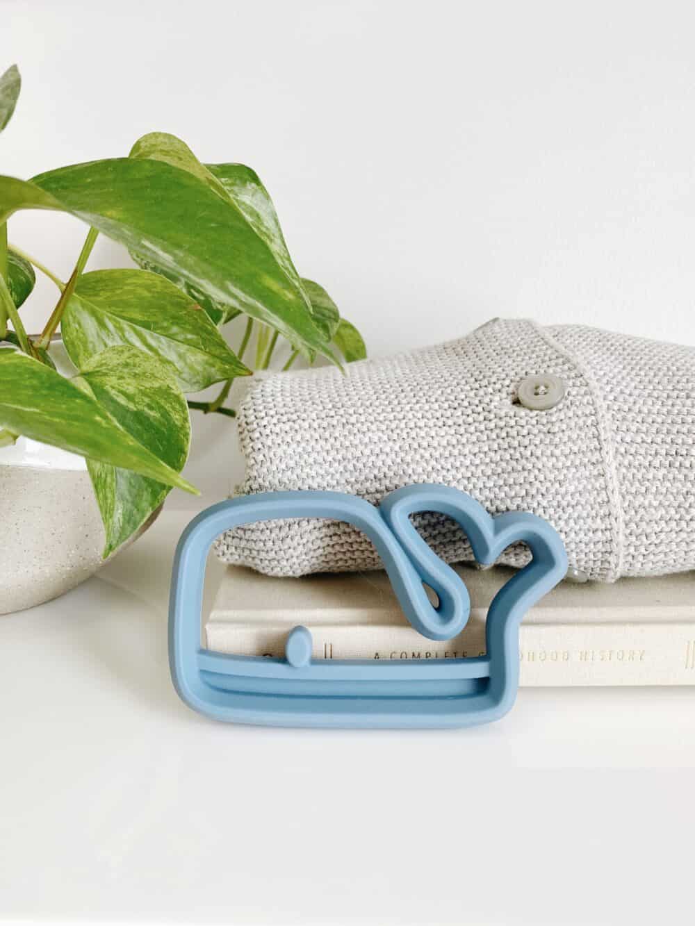 A blue whale shaped teether on a shelf next to a plant.