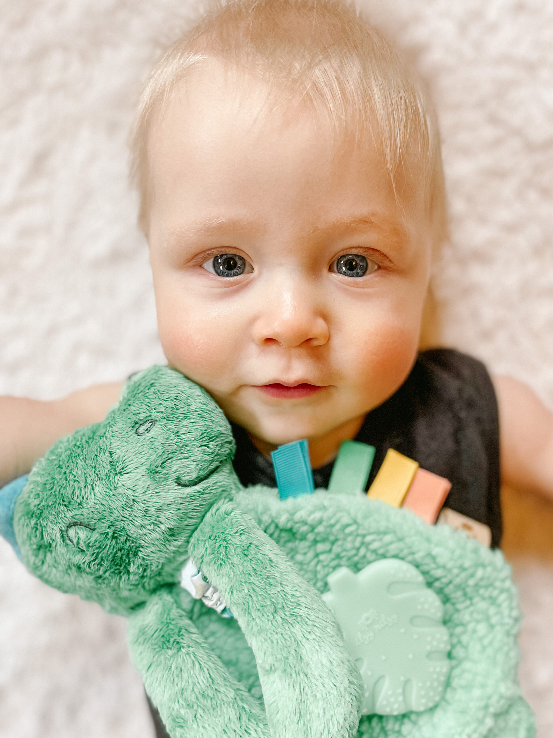 A baby with light hair lies on a white blanket, holding a green plush toy. The baby is wearing a dark outfit and looking directly at the camera.