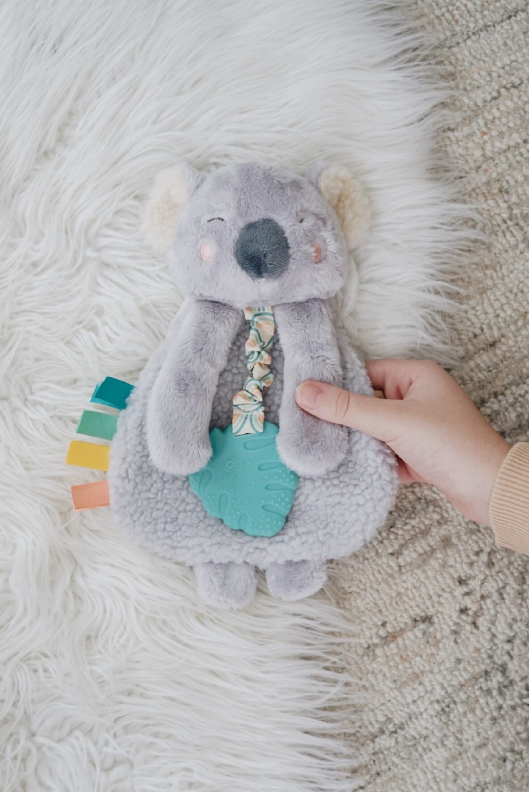 A hand holding a plush koala toy with a teal teether attached to its front and colorful fabric tabs on its side, lying on a fluffy white surface.