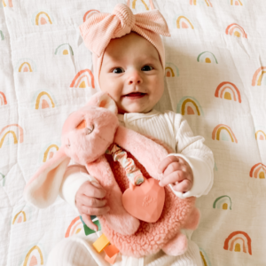 A baby dressed in white with a pink headband smiles while holding a pink stuffed animal. The background features a white blanket with colorful rainbow patterns.
