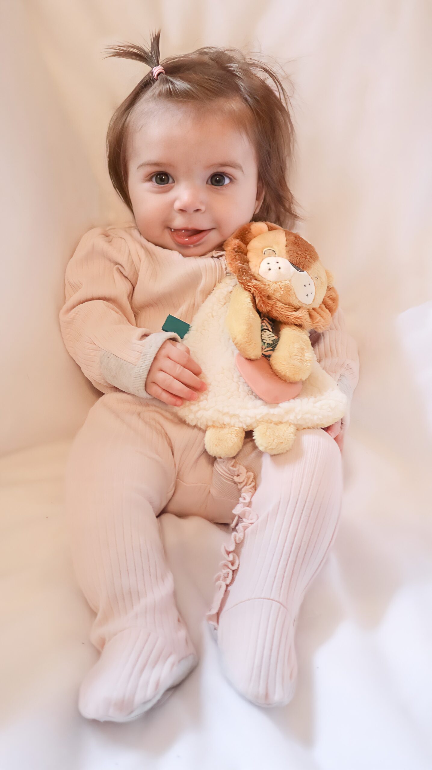 A baby in light pink clothing sits on a white surface, holding a stuffed lion toy.