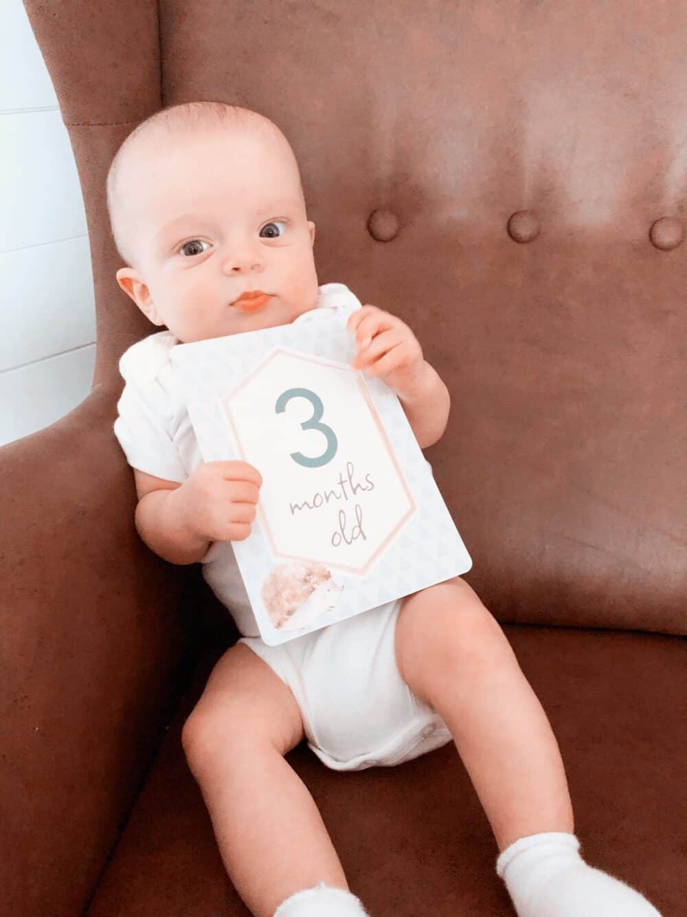 A baby is sitting in a brown chair holding a number sign.