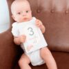 A baby is sitting in a brown chair holding a number sign.