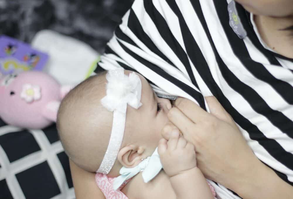 A baby is being breastfed by a woman wearing a Mom Boss™ 4-IN-1 Multi-Use Nursing Cover & Scarf.