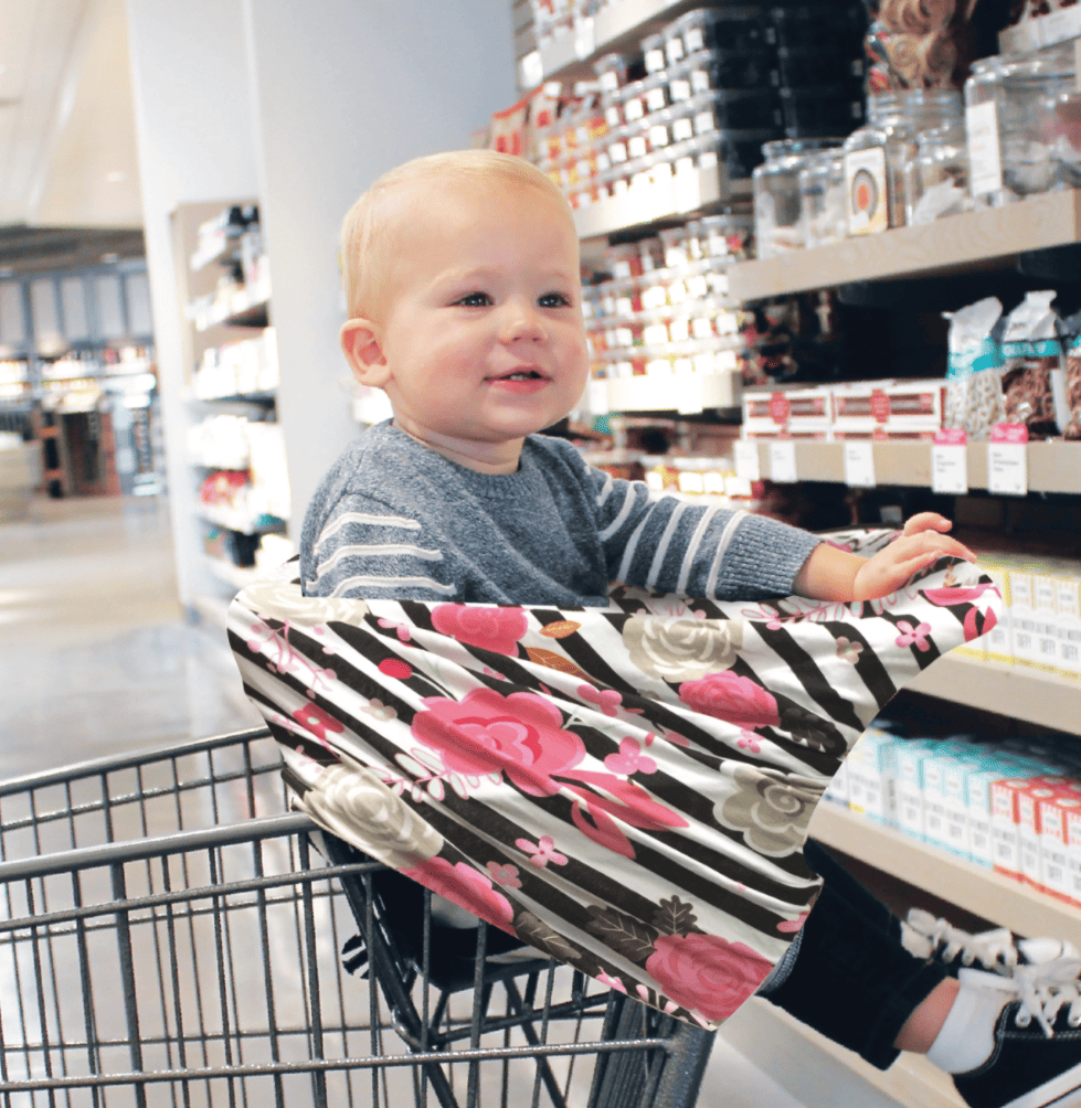 A baby sitting in a Mom Boss™ 4-IN-1 Multi-Use Nursing Cover & Scarf in a grocery store.