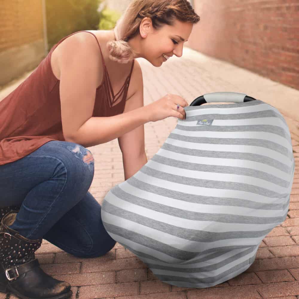 A woman is kneeling down on a sidewalk with a Mom Boss™ 4-IN-1 Multi-Use Nursing Cover & Scarf car seat cover.