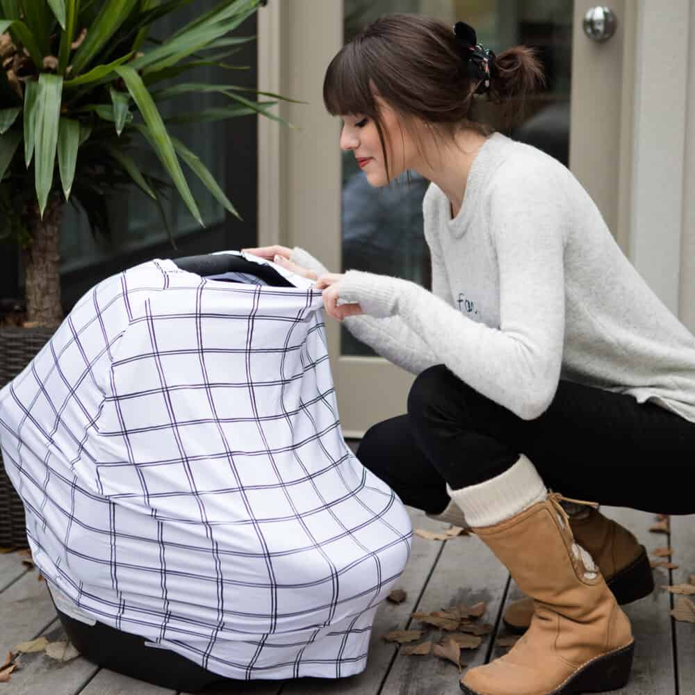 A woman kneeling in front of a Mom Boss™ 4-IN-1 Multi-Use Nursing Cover & Scarf with a plaid cover.